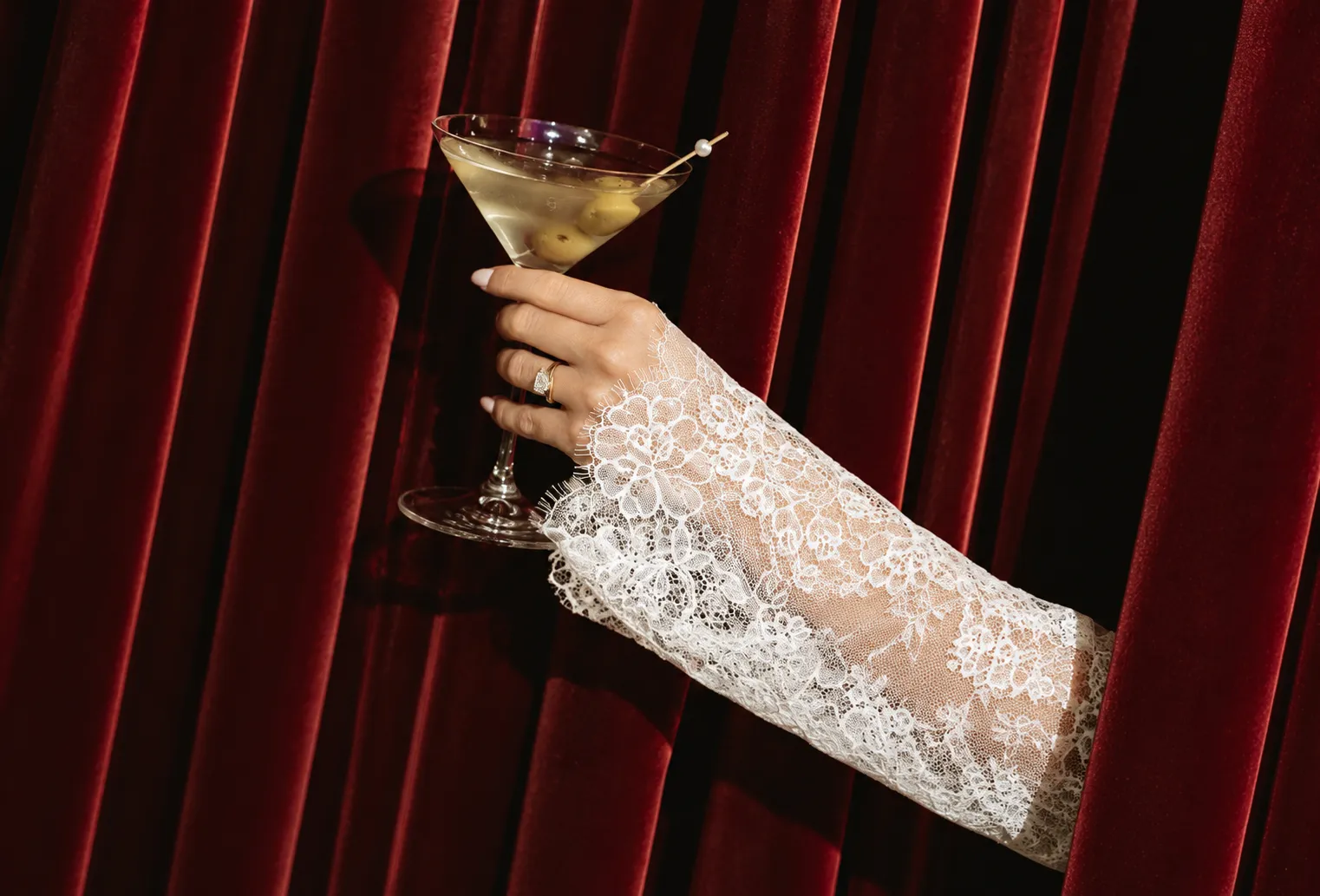 A lace-sleeved hand holding a martini glass out from behind a red velvet curtain