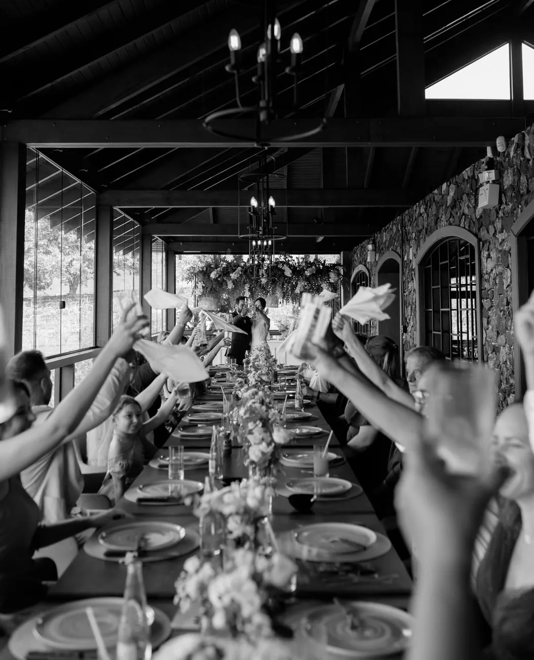 Black-and-white photo of wedding guests at a long table, raising their napkins in celebration