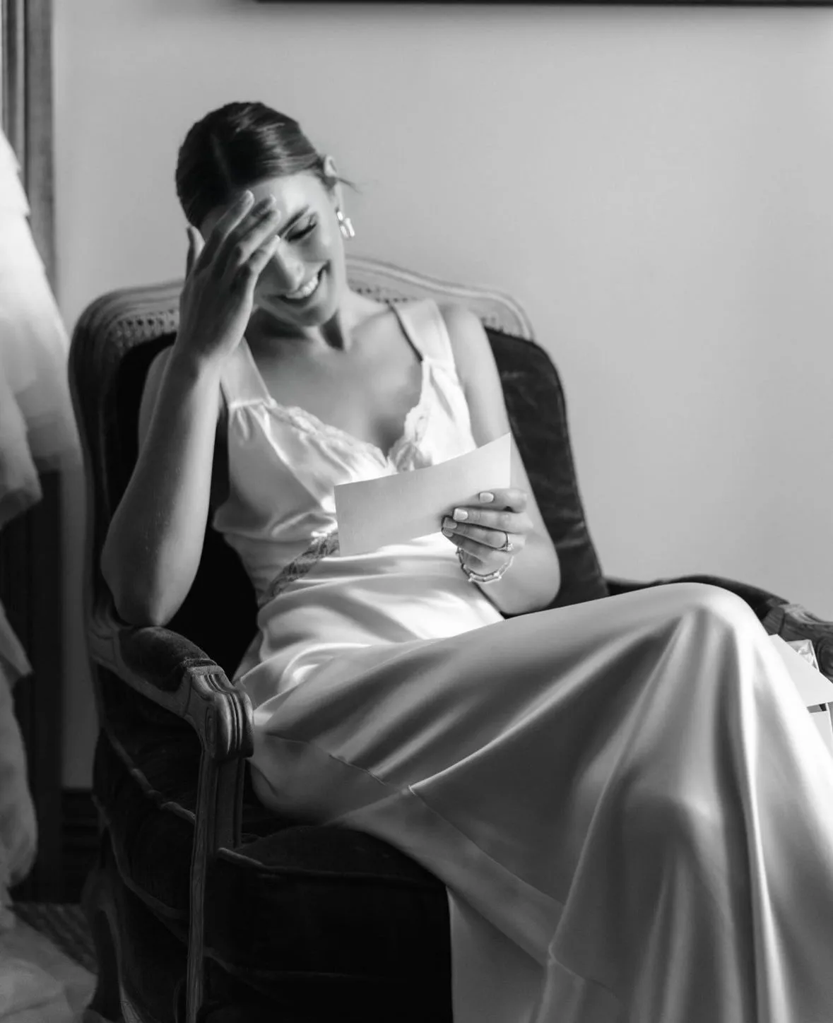 A bride in a satin dress laughing as she reads a letter
